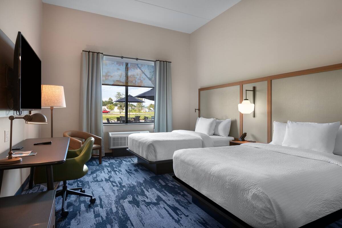 Quarto de hotel com duas camas, uma escrivaninha, uma cadeira, uma televisão e uma janela com vista para o exterior.