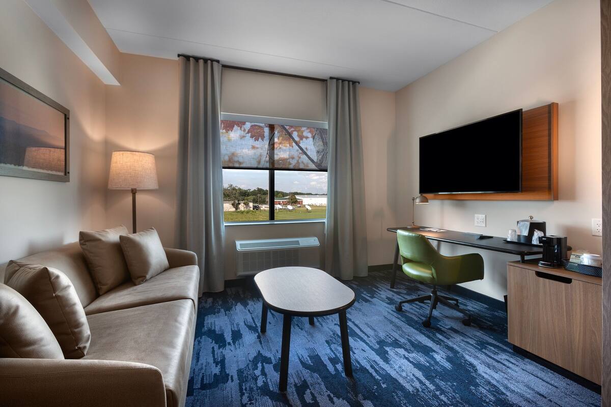 Quarto de hotel com sofá, escrivaninha, cadeira, televisão e janela com vista para o exterior.