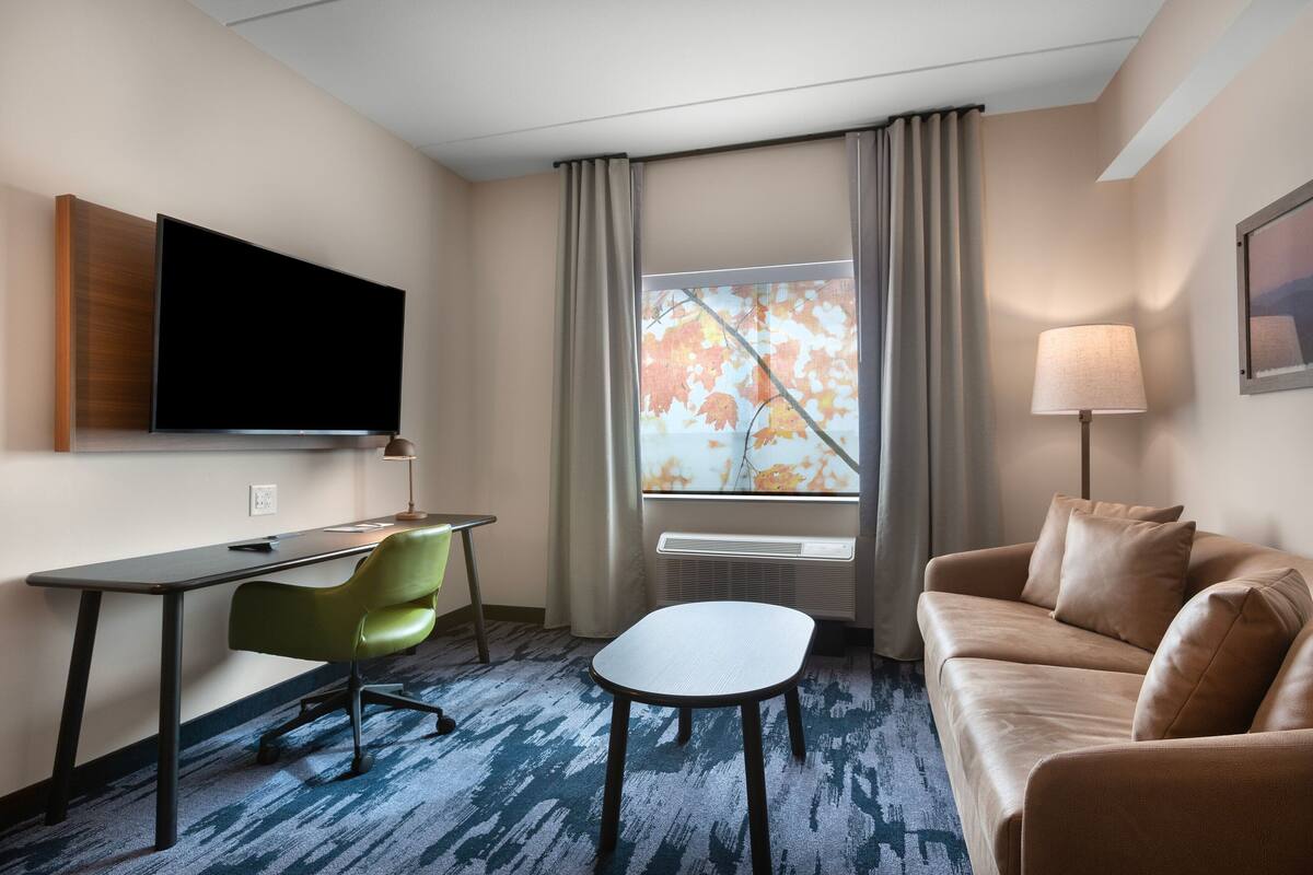 Quarto de hotel com televisão de tela plana na parede, uma escrivaninha com cadeira, sofá, mesa pequena e janela com cortinas.