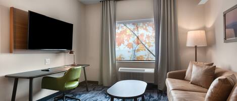 A hotel room with a flat-screen TV mounted on the wall, a desk with a chair, a sofa, a small table, and a window with curtains.