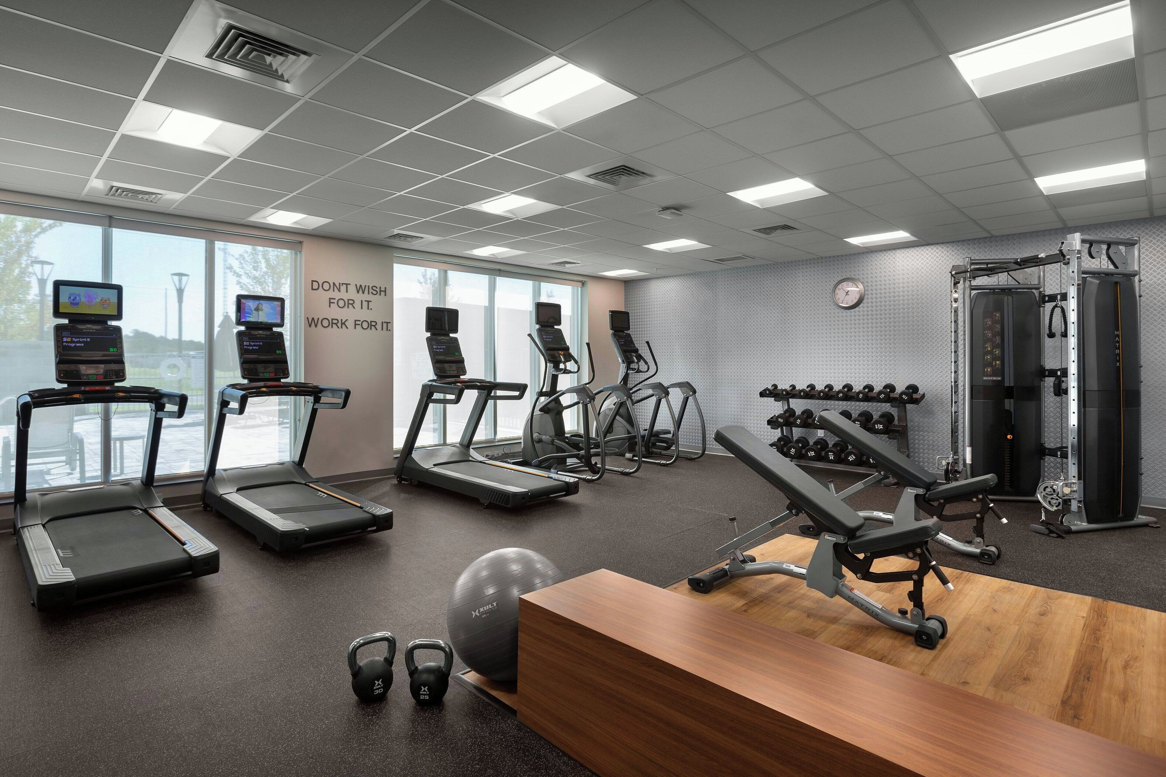 A modern gym with treadmills, elliptical machines, free weights, and a wooden reception desk.