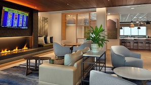 A modern lounge area with a fireplace, seating, a television displaying sports content, and a kitchen area in the background. - Fairfield Inn & Suites By Marriott Coastal Carolina Conway (Conway)