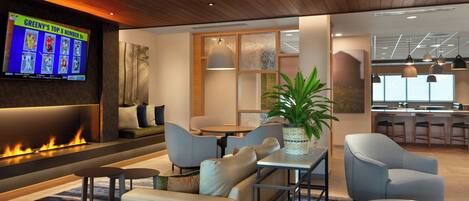 A modern lounge area with a fireplace, seating, a television displaying sports content, and a kitchen area in the background.