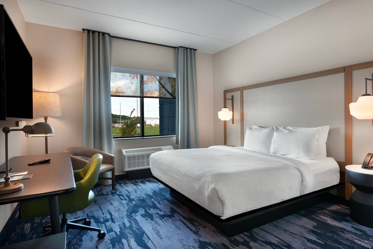 Quarto de hotel com uma cama grande, uma escrivaninha com cadeira, televisão e janela com cortinas.