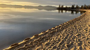 En strand med fodspor, der fører ud i roligt vand, omgivet af træer og fjerne bjerge.