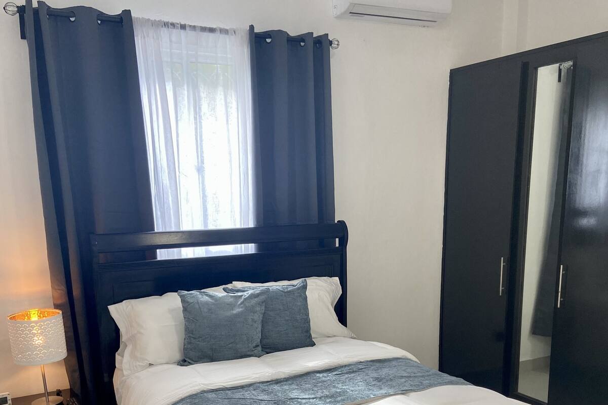 Una cama bien hecha con edredón blanco y gris, una lámpara de noche y una mesita de noche de madera oscura.