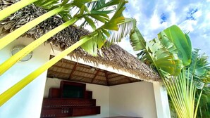 Uma área de estar coberta ao ar livre com cadeiras de madeira e um bar, cercada por plantas tropicais e uma estrutura de telhado de palha.
