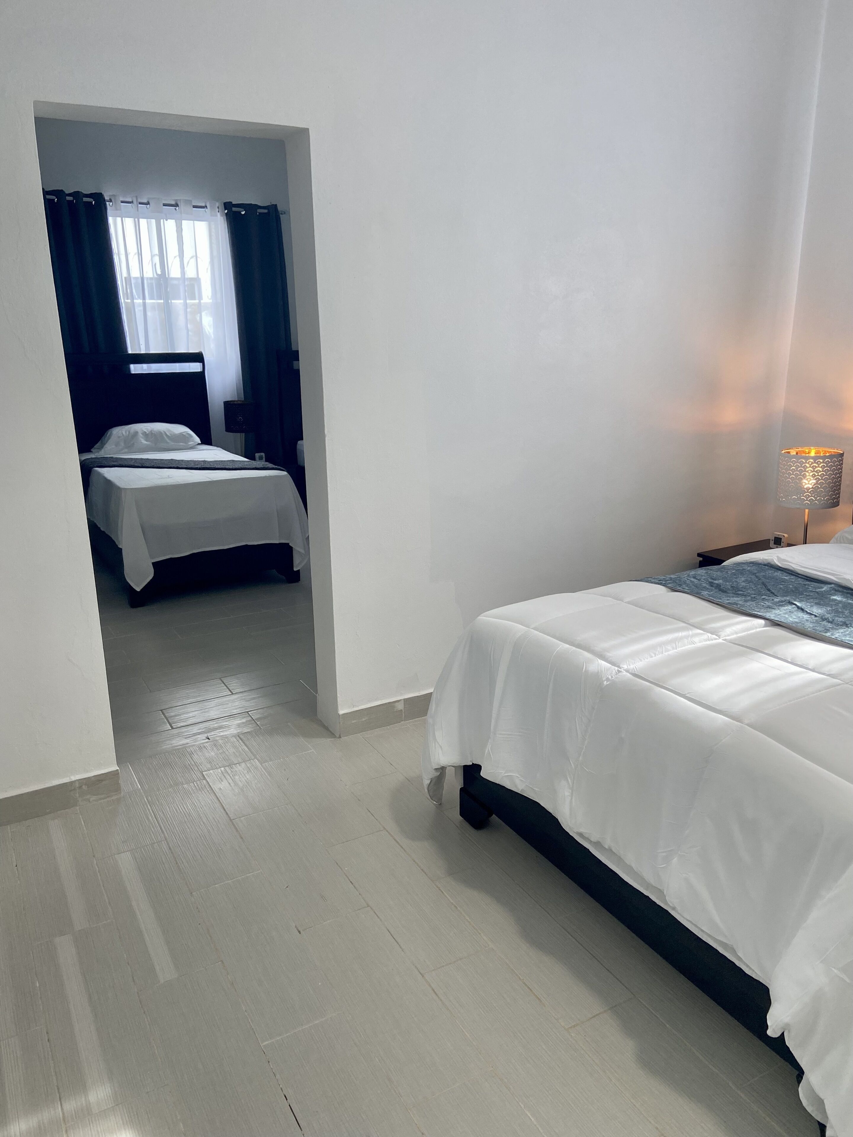 A hotel room with a bed, a nightstand with a lamp, and a view through an open doorway to another room.