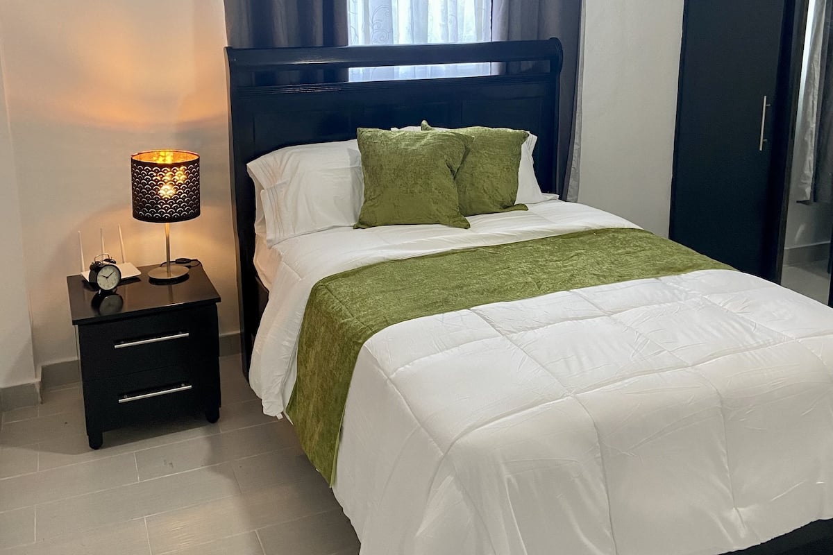 Una cama bien tendida con ropa blanca y verde, una lámpara de noche y una mesita de noche con un reloj.
