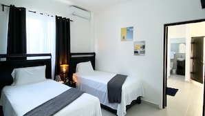 Quarto de hotel com duas camas de solteiro, uma mesa de cabeceira com abajur, ar-condicionado no teto e vista para o banheiro por uma porta aberta.