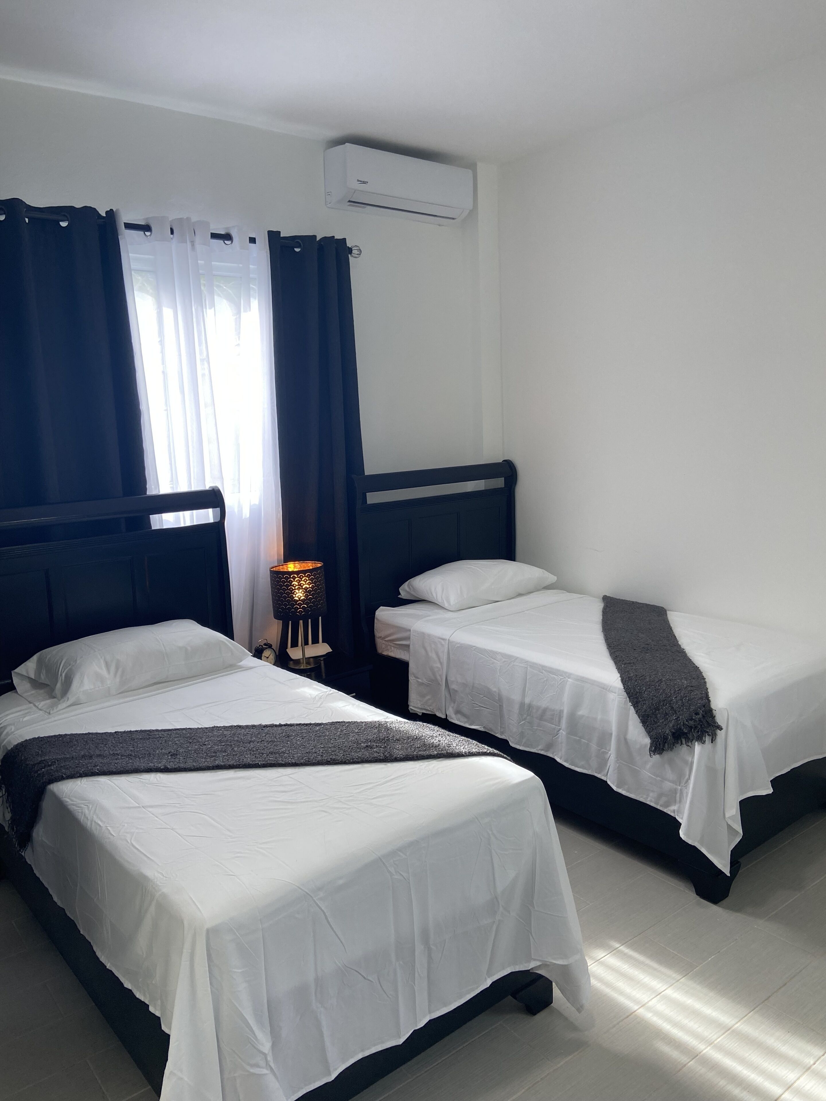 A hotel room with two single beds, a nightstand with a lamp, and an air conditioning unit on the wall.