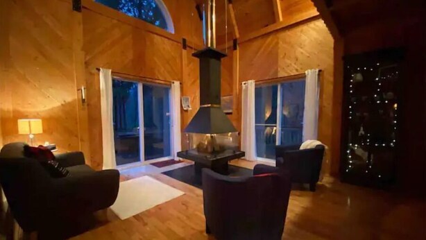 A cozy wooden interior with a fireplace, two sofas, and a large window. - Cottage by River Falls with Hot Tub & Sauna (Amherst)