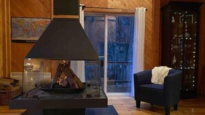 A cozy room with a wood-burning stove, a dark blue armchair, and a glass door leading to a balcony with a view of trees. - Cottage by River Falls with Hot Tub & Sauna (Amherst)