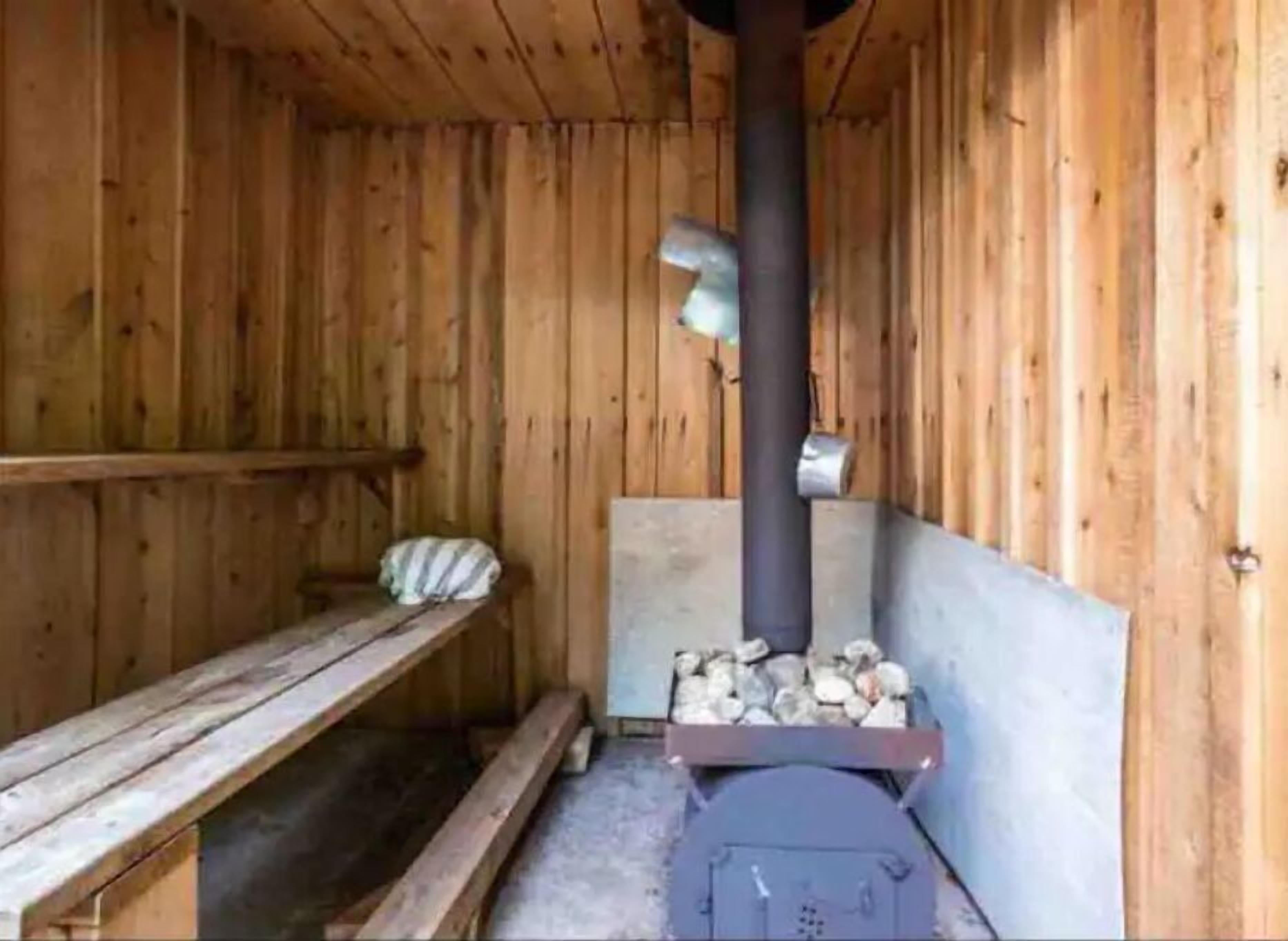 A wooden sauna room with a bench, a wood-burning stove, and a bench with a pillow.