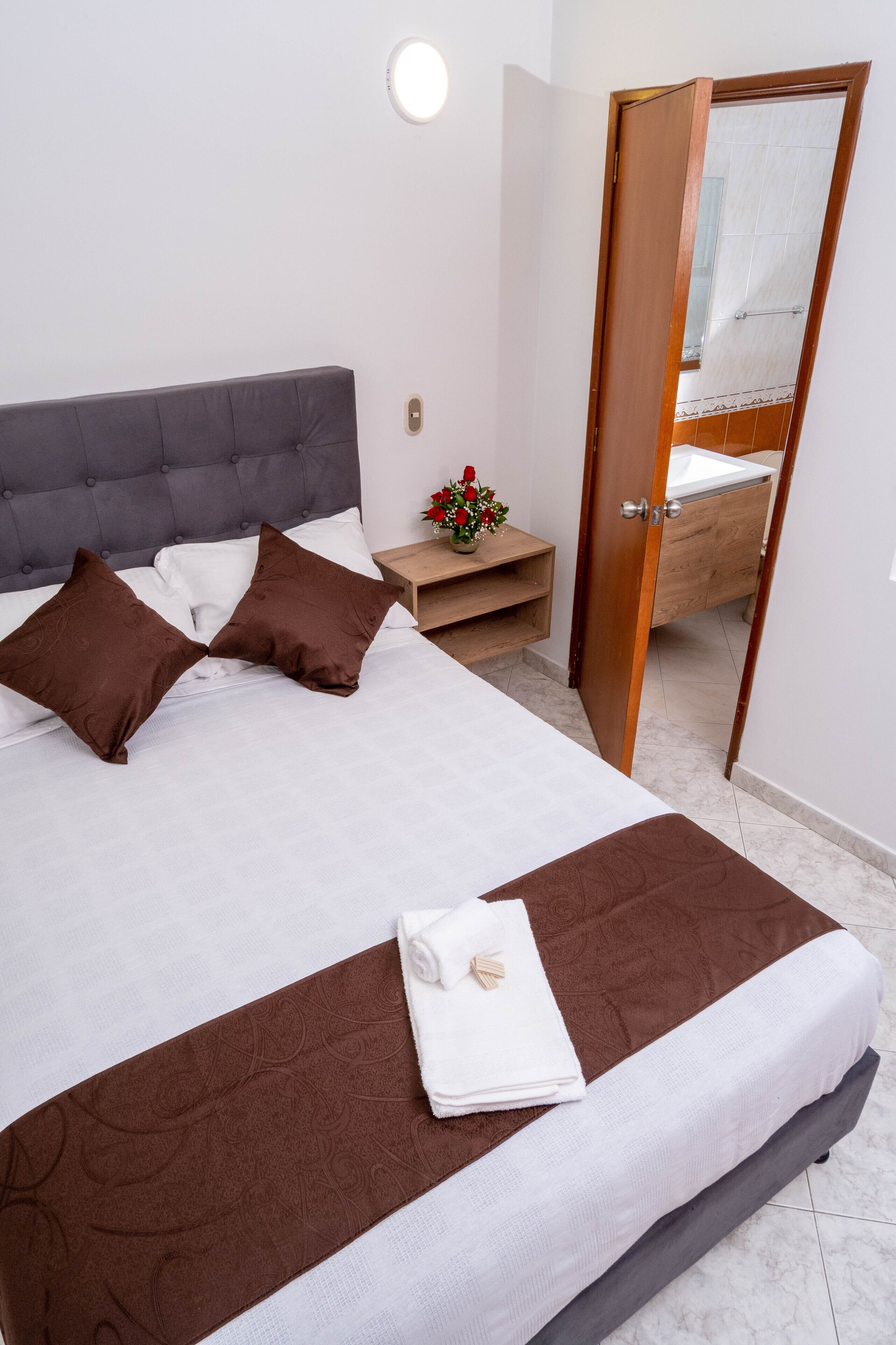 Un lit bien fait, avec des oreillers marron, une couette blanche et un couvre-lit marron. On aperçoit une table de chevet en bois sur laquelle est posé un vase de fleurs, ainsi qu’une petite table. Une salle de bain est visible à travers une porte ouverte.