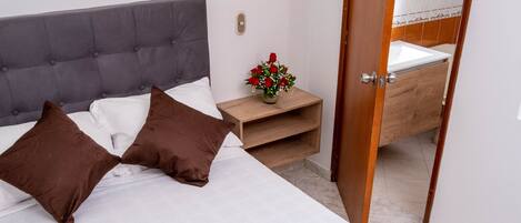Un lit bien fait, avec des oreillers marron, une couette blanche et un couvre-lit marron. On aperçoit une table de chevet en bois sur laquelle est posé un vase de fleurs, ainsi qu’une petite table. Une salle de bain est visible à travers une porte ouverte.