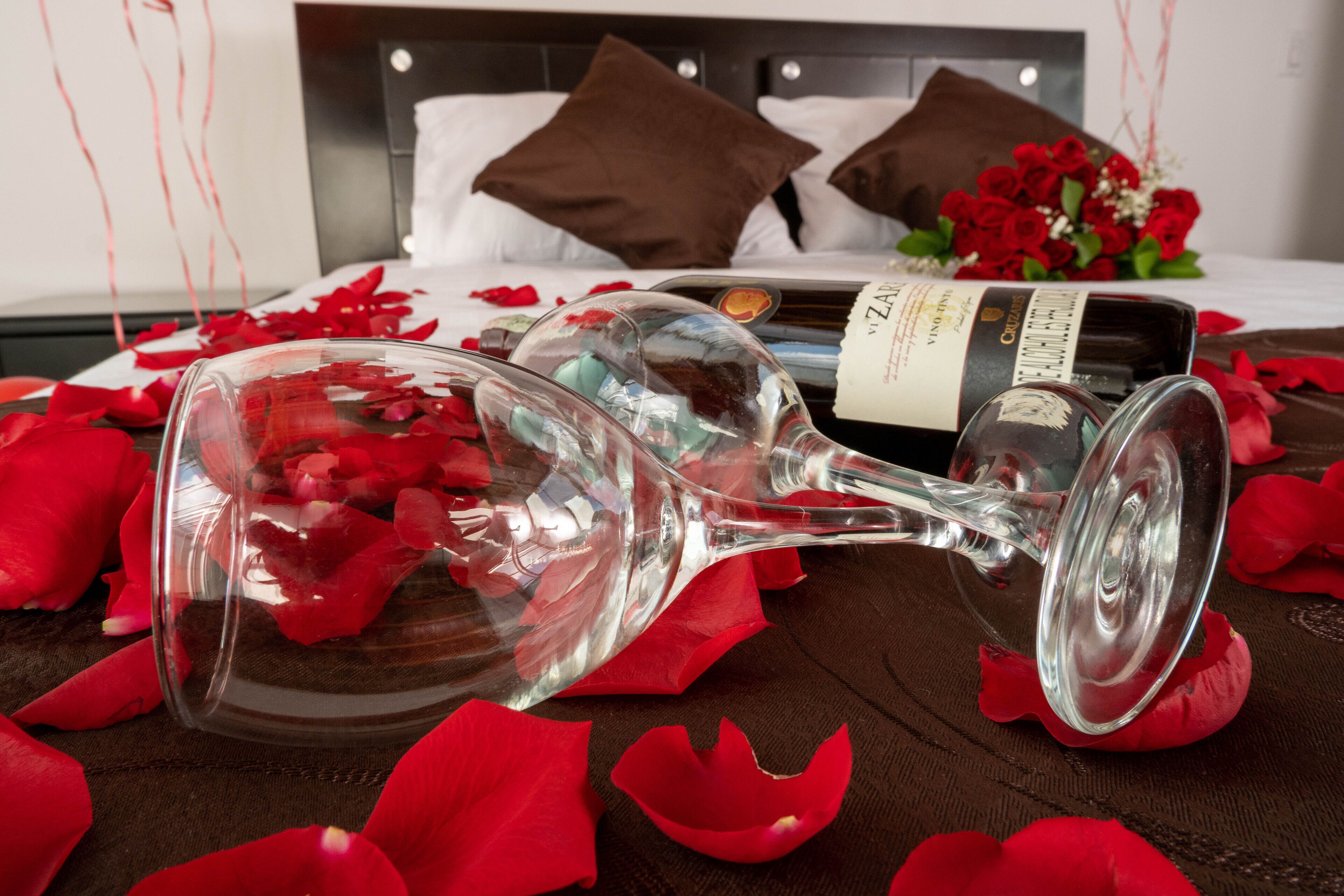 Ein Bett mit weißer Decke, zwei braune Kissen, ein Rosenstrauß, eine Weinflasche und verstreute Rosenblätter.