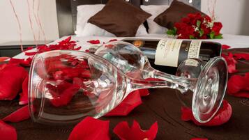 Ein Bett mit weißer Decke, zwei braune Kissen, ein Rosenstrauß, eine Weinflasche und verstreute Rosenblätter.