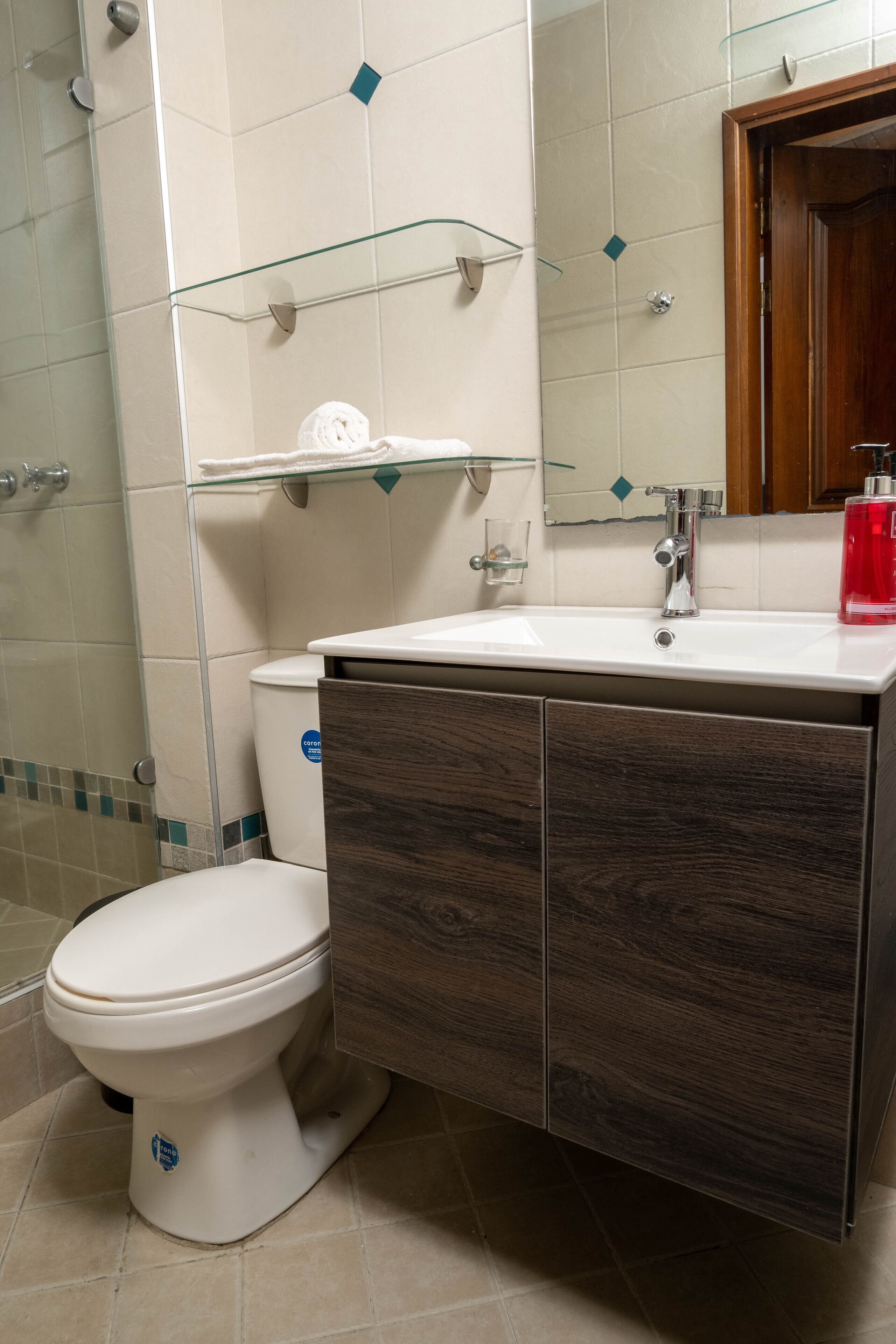 A bathroom with a white toilet, a wooden cabinet, a glass shelf, and a mirror above the sink.