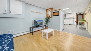 A room with a bed, a TV, a small table, and a kitchen area in the background.
