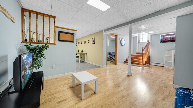 A spacious room with wooden flooring, a white table, a TV on a stand, a plant, and a staircase leading to another area.