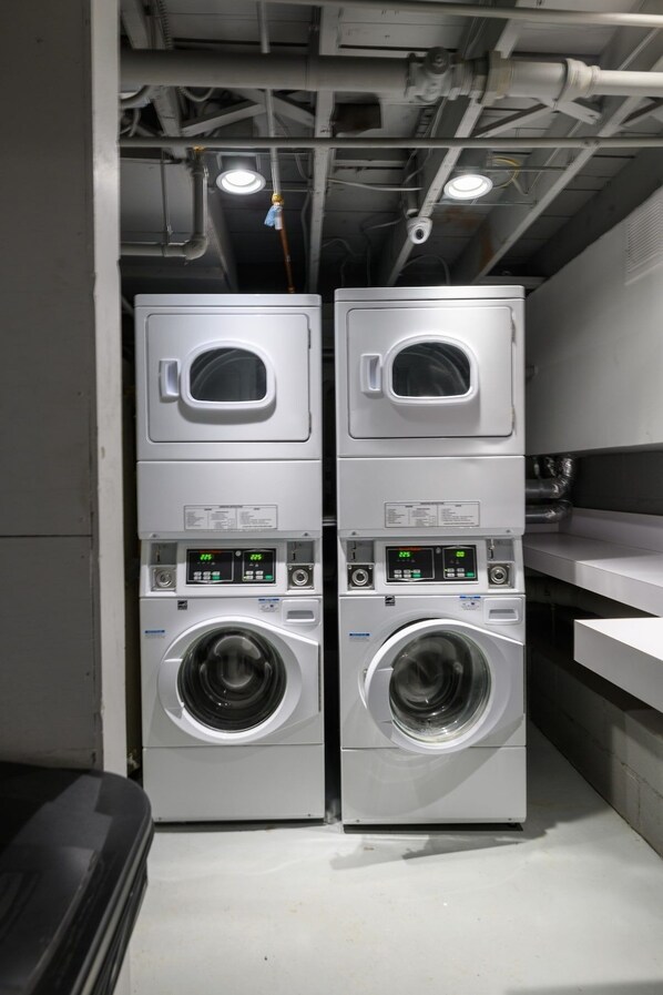 Deux machines à laver à chargement frontal, superposées, dans une buanderie, avec des panneaux de commande visibles et un plafond où les tuyaux et les luminaires sont apparents.