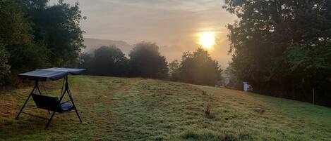 Ein grasbewachsenes Feld mit einer Camping-Ausrüstung, darunter ein Tisch und Stühle, umgeben von Bäumen und einem entfernten Blick auf den untergehenden Sonnenuntergang.