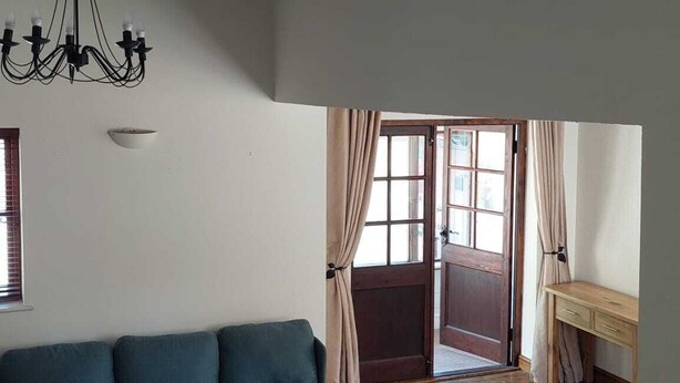 Ein Wohnzimmer mit einem blauen Sofa, einem Holztisch und einem Streifen Teppich. Es gibt eine Chandelüster, ein Fenster mit Vorhängen und eine Holzbank.