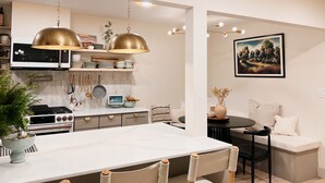 Une cuisine moderne dotée d’un plan de travail blanc, d’appareils électroménagers en acier inoxydable et de luminaires suspendus. On y trouve également un coin repas avec une table et des chaises blanches.