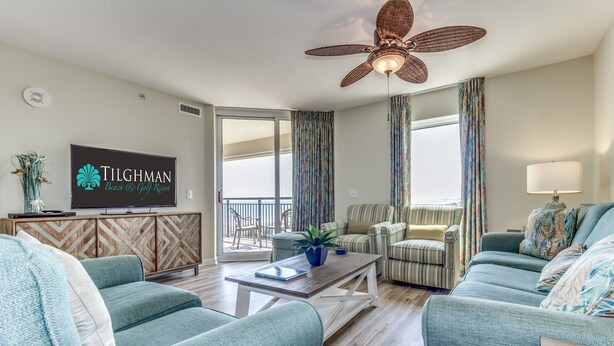 Ein geräumiges Wohnzimmer mit türkisfarbenen Sofas, einem Holztisch und einem Deckenventilator. Ein Fernseher ist an der Wand montiert, und durch große Fenster ist der Balkon zu sehen.