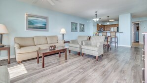A spacious living room with a sofa, armchairs, a coffee table, and a ceiling fan. A kitchen is visible in the background with bar stools.
