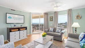 Amplia sala de estar con un juego de sofás, una mesa de centro con flores, un televisor montado en la pared y vistas al océano a través de grandes ventanales.