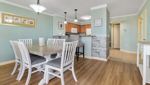 Un comedor con una mesa de madera y sillas blancas, una cocina con armarios de madera y un pasillo que lleva a otra habitación.