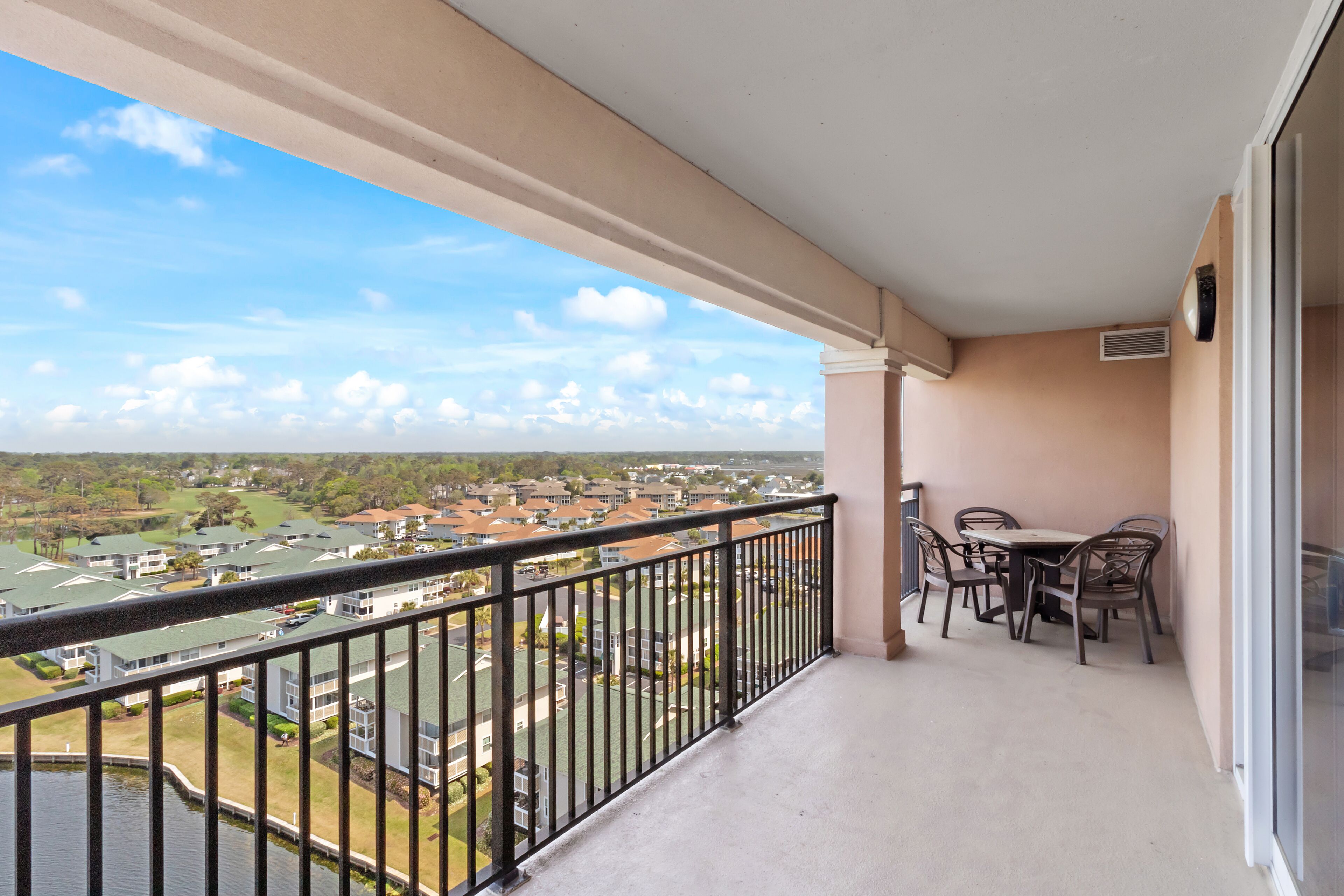 A balcony with a table and chairs, overlooking a residential area with houses and a body of water.