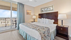 Une chambre d’hôtel avec un grand lit, deux tables de chevet avec des lampes, un ventilateur de plafond, un balcon avec vue et des œuvres d’art au mur.