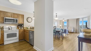 A modern kitchen with wooden cabinets, a microwave, stove, and dishwasher. There is a dining area with a table and chairs, and a view of the city through large windows. - 3BR- 5th floor- North Lake & Oceanview Back Corner|Free Parking| Resort Access| (North Myrtle Beach)