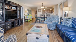 Un salon avec un canapé, une table basse et un téléviseur. On y trouve également un coin repas avec une table et des chaises. La cuisine est visible au fond.