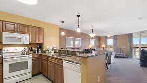 A modern kitchen with a built-in oven, microwave, and dishwasher. There is a large island with a sink and a dining area with a chair and table. The room has large windows and a view of the outdoors.