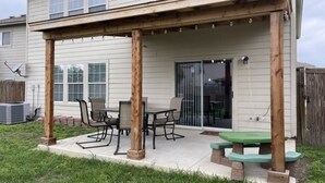 Eine überdachte Terrasse mit Tisch und Stühlen, eine Holz-Pergola und ein grüner Picknicktisch.