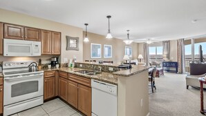 A modern kitchen with wooden cabinets, a built-in microwave, and a dishwasher. There are pendant lights above the counter and a large island with a sink. The kitchen leads to a dining area with a view of the mountains. - 3BR- 14th floor- Lake & Golf View condo|Short walk to beach| Resort Access| (North Myrtle Beach)