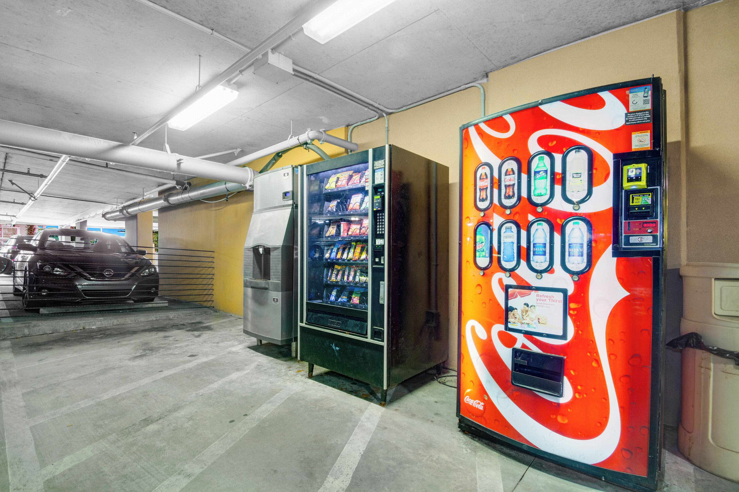 Un parking avec des distributeurs automatiques, dont un aux couleurs de Coca-Cola, et une voiture en arrière-plan.