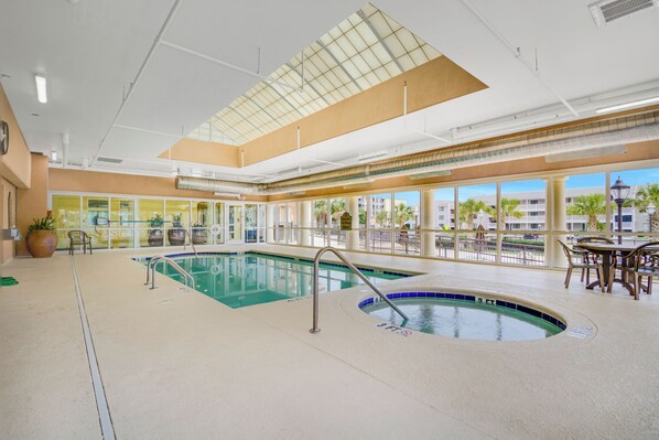 Une piscine intérieure avec des parois en verre transparent, un toit ouvrant et un espace détente avec des tables et des chaises.