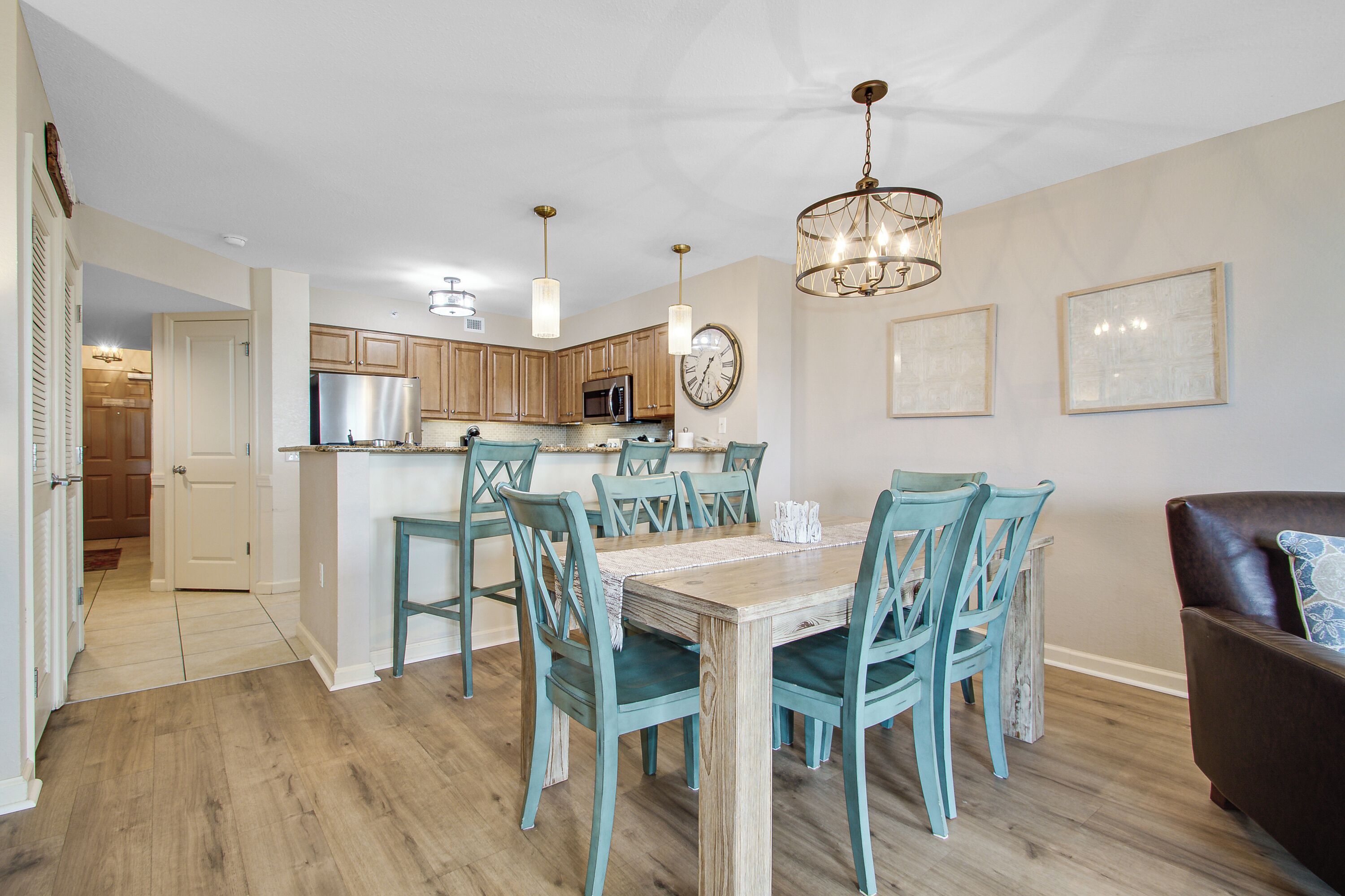 A modern kitchen with wooden cabinets, a stainless steel refrigerator, and a dining area with a wooden table and teal chairs.