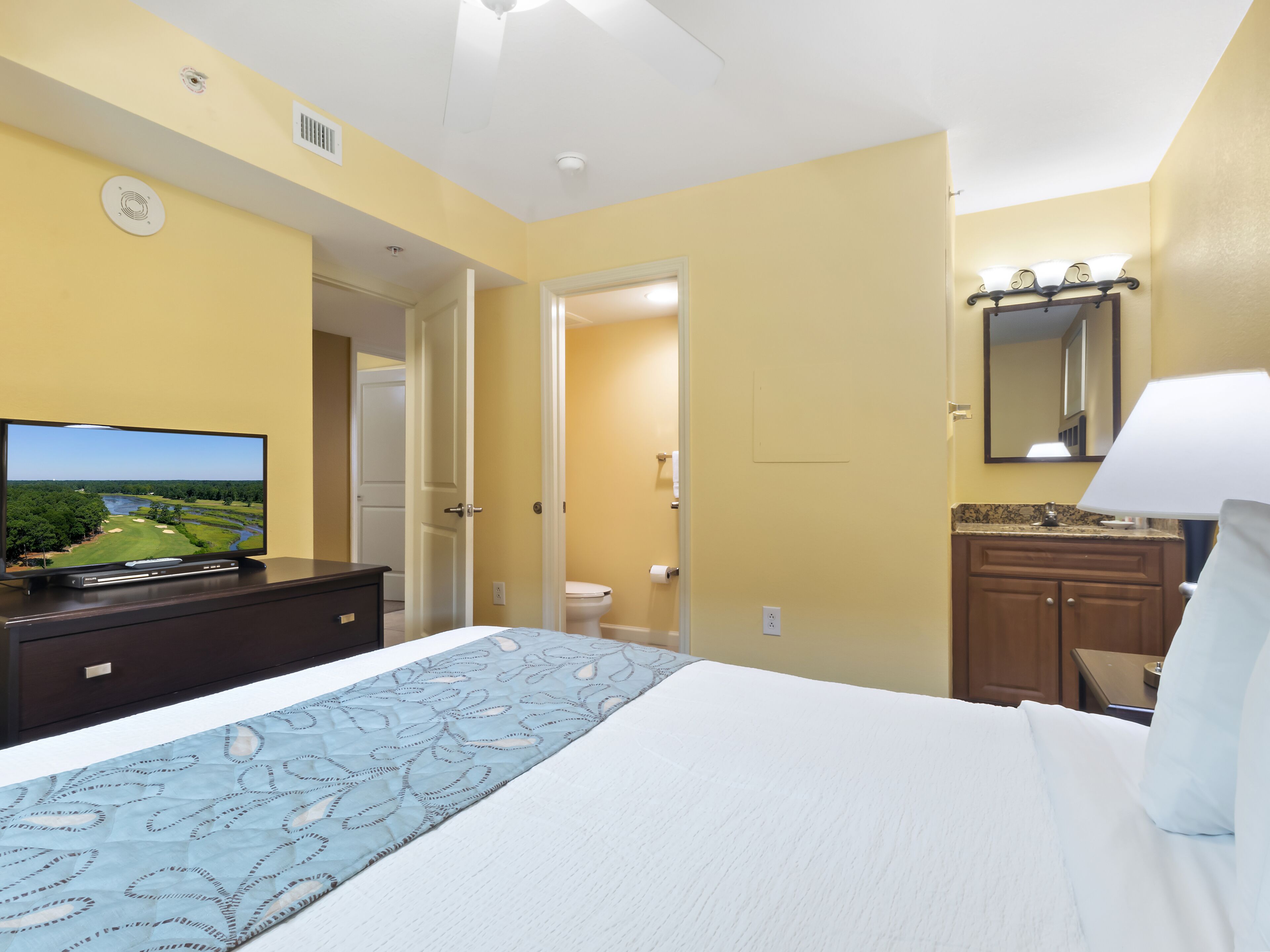 A hotel room with a bed, a TV on a dresser, a bathroom visible through an open door, and a mirror above the sink.