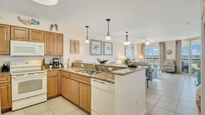 A modern kitchen with wooden cabinets, a white microwave, and a dishwasher. There are two pendant lights, a coffee maker, and a view of the balcony with a chair and table.