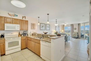 A modern kitchen with wooden cabinets, a white microwave, and a dishwasher. There are two pendant lights, a coffee maker, and a view of the balcony with a chair and table.