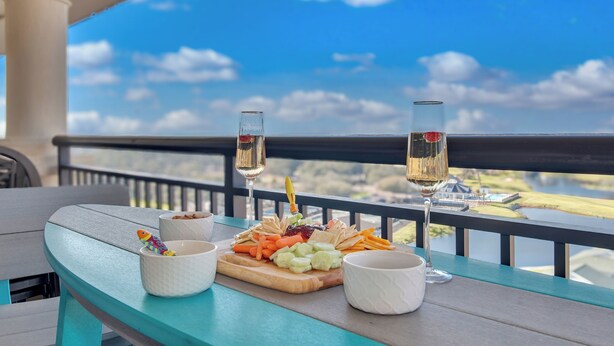 A table with a fruit and vegetable platter, two glasses of white wine, and a small decorative fish on a balcony with a scenic view.