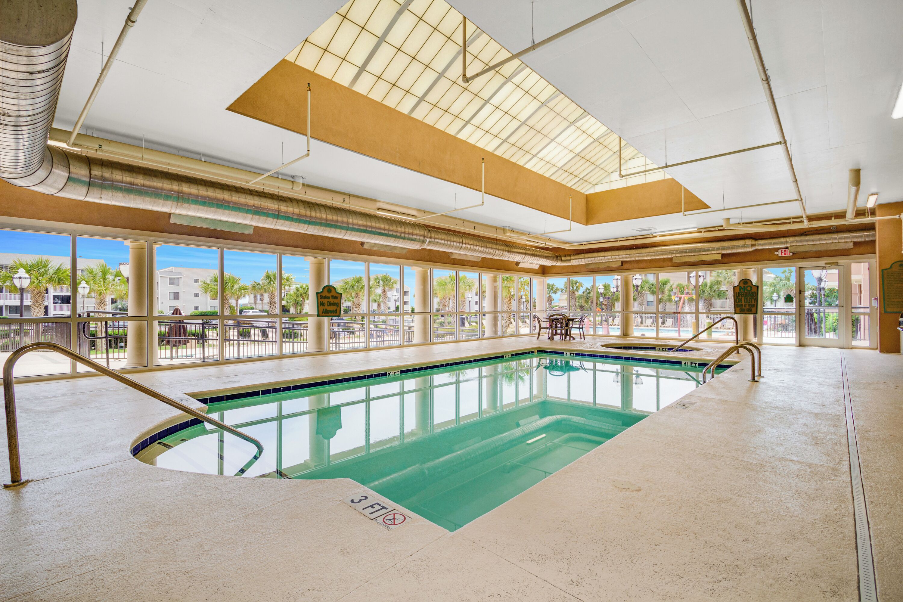 Une piscine intérieure avec des parois en verre transparent, une verrière et une vue sur les palmiers à l’extérieur.
