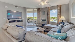 A modern living room with a flat-screen TV, comfortable seating, a ceiling fan, and a view of the outdoors. - 3BR- 7th floor- Lake & Golf View condo|Short walk to beach| Free parking (North Myrtle Beach)