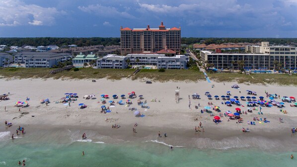 A beach with numerous umbrellas and people. There are buildings in the background.